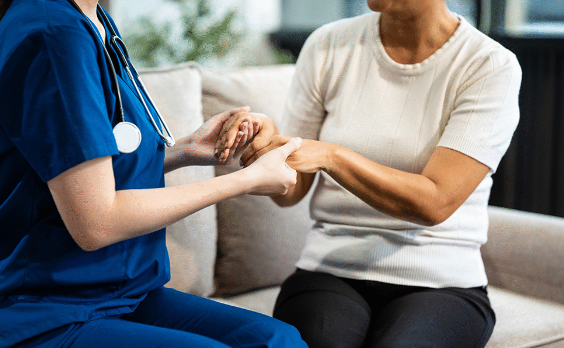 Une jeune infirmière asiatique, seule femme présente, tient la main d'une femme d'âge mûr aux cheveux gris, assise sur un canapé, et l'encourage tout en recueillant ses antécédents médicaux et en procédant à des examens de santé.