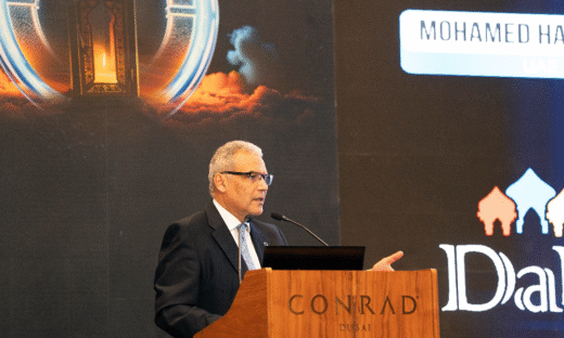 Prof Mohamed Hassanein stands at a podium. He is wearing a dark suit and tie and speaks into a microphone while gesturing with one hand. Behind him, a large event screen displays vibrant graphics, including an illuminated lantern motif and text elements, alongside the logo of the DaR programme.